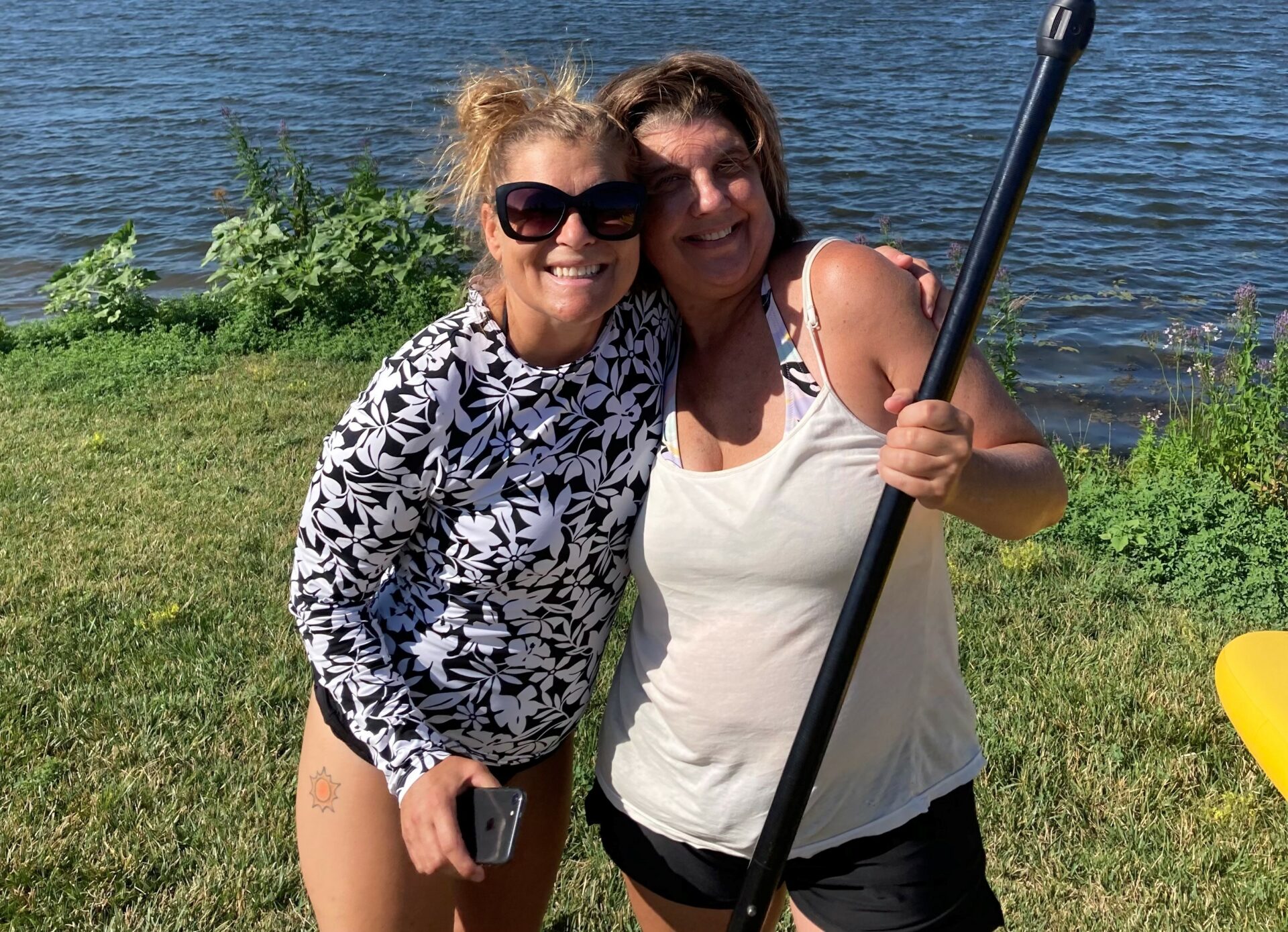 Two ladies standing side by side in bathing suits and tanks tops. One is holding an oar and they are in front of a body of water.