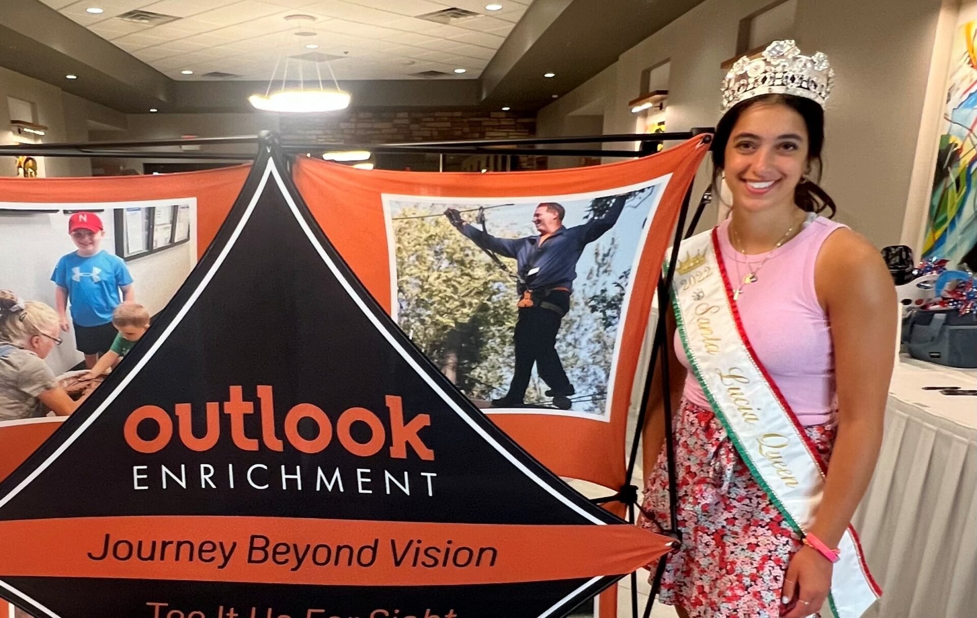 The 2022 Santa Lucia Italian Queen standing next to an Outlook Enrichment sign. She is a beautiful, smiling young lady with dark hair and is wearing a crown.