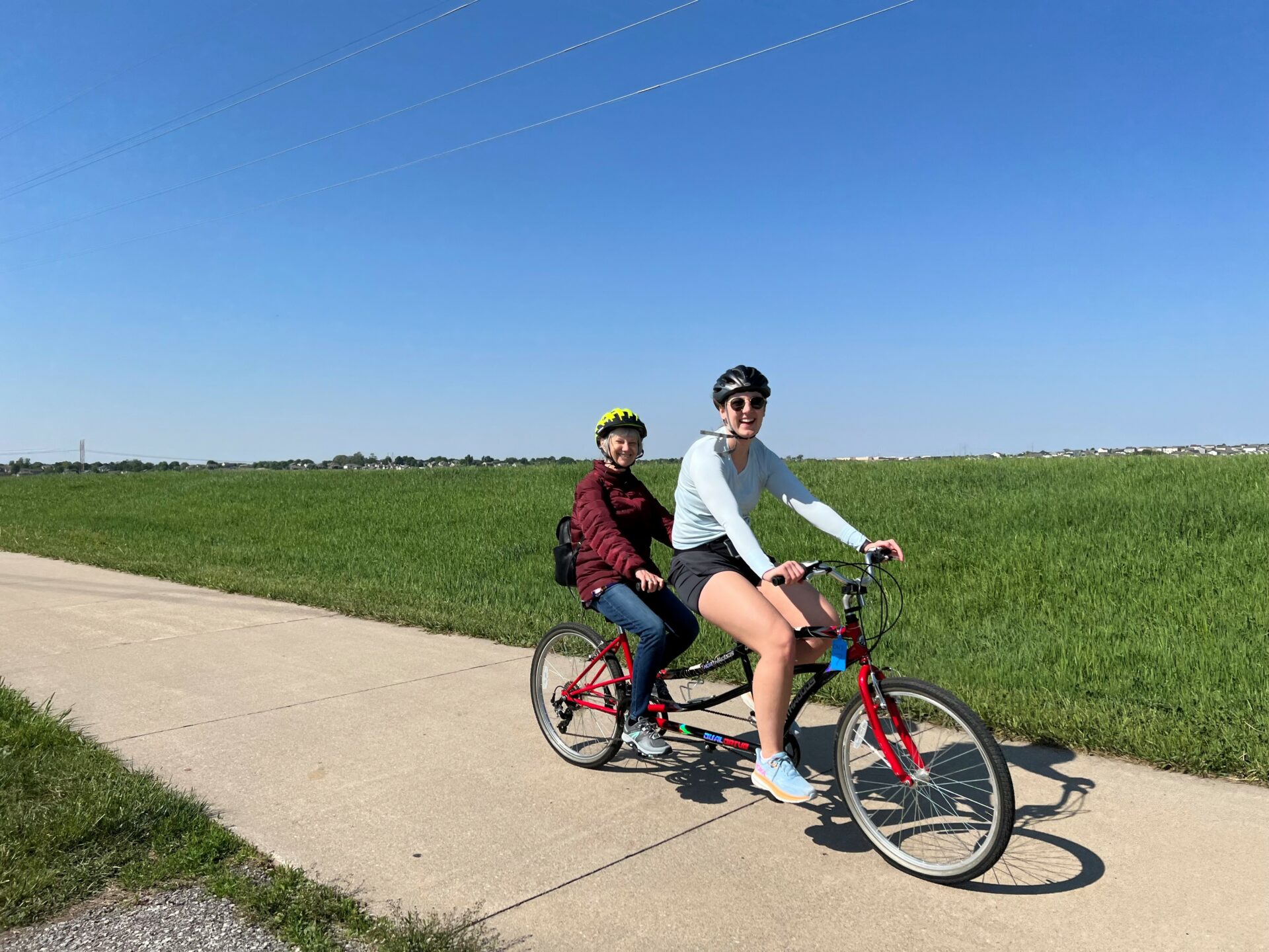 Two ladies on a tandem bike wearing helmets. The one in front is college aged and the one in back is retired. They are both smiling and on a trail with green grass in the background.