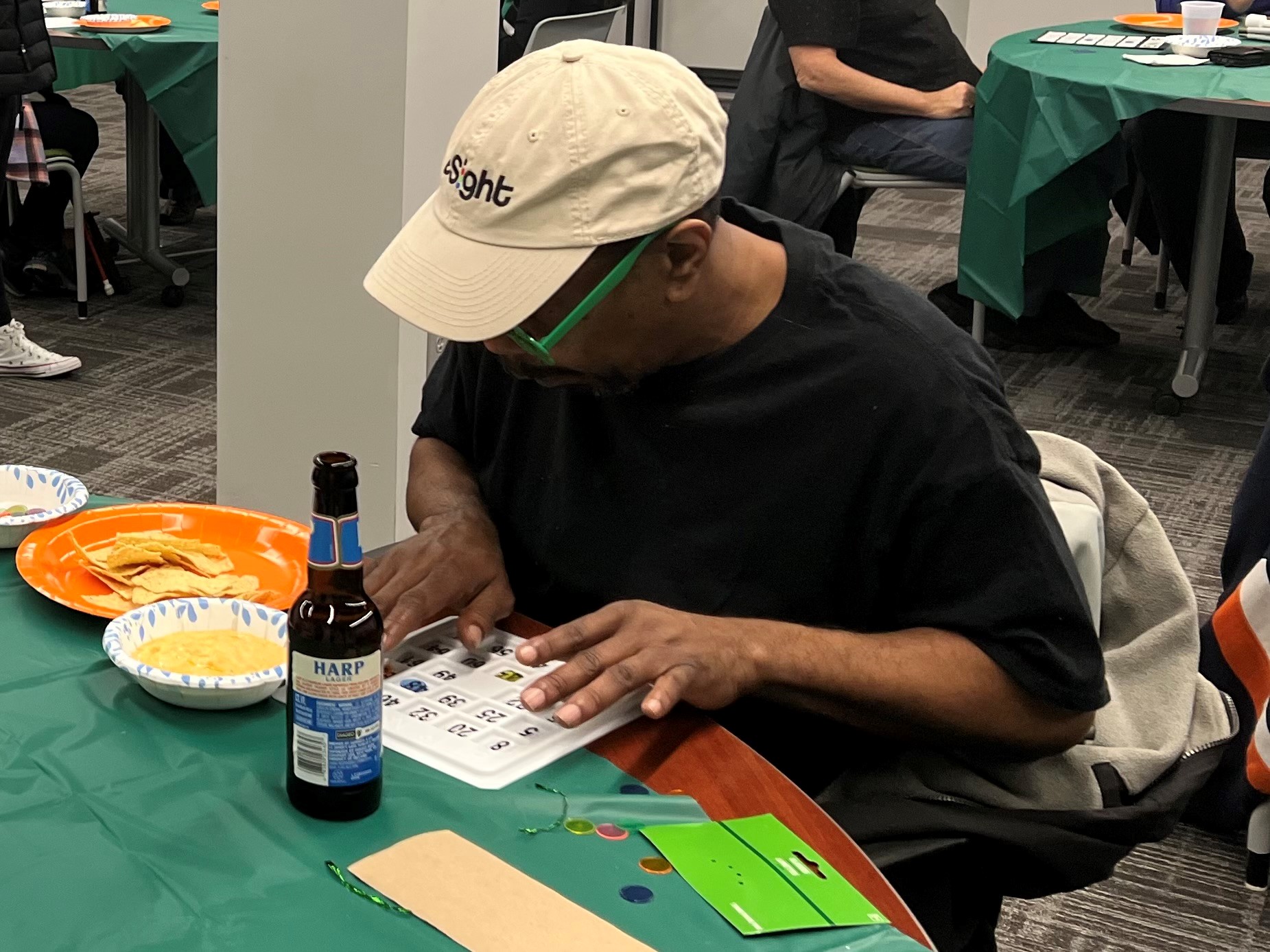 A black man with a hat on sitting at a table. His fingers are reading the letters and numbers on his Bingo card.