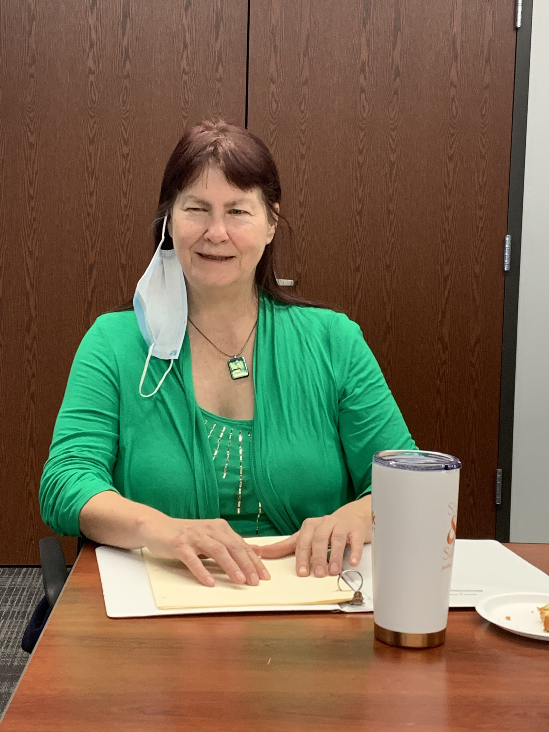 A woman with long hair and a green shirt is sitting at a table. She has a poem she wrote in Braille in front of her. Her hands are on the paper and she is reading the poem out loud.