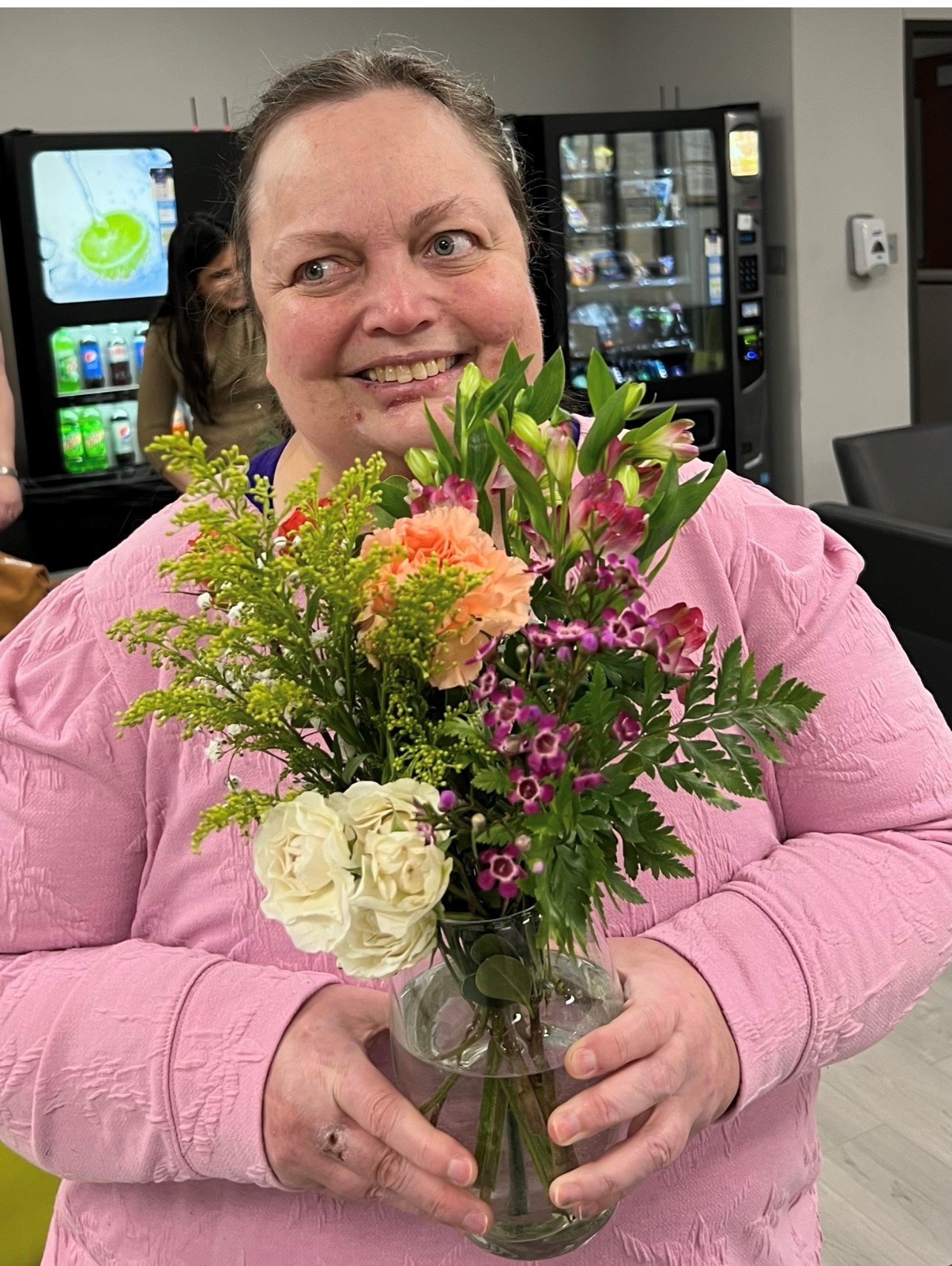 A woman is sitting at a table with a big smile on her face. She is holding a vase full of fresh flowers so big that she can barely see over it.