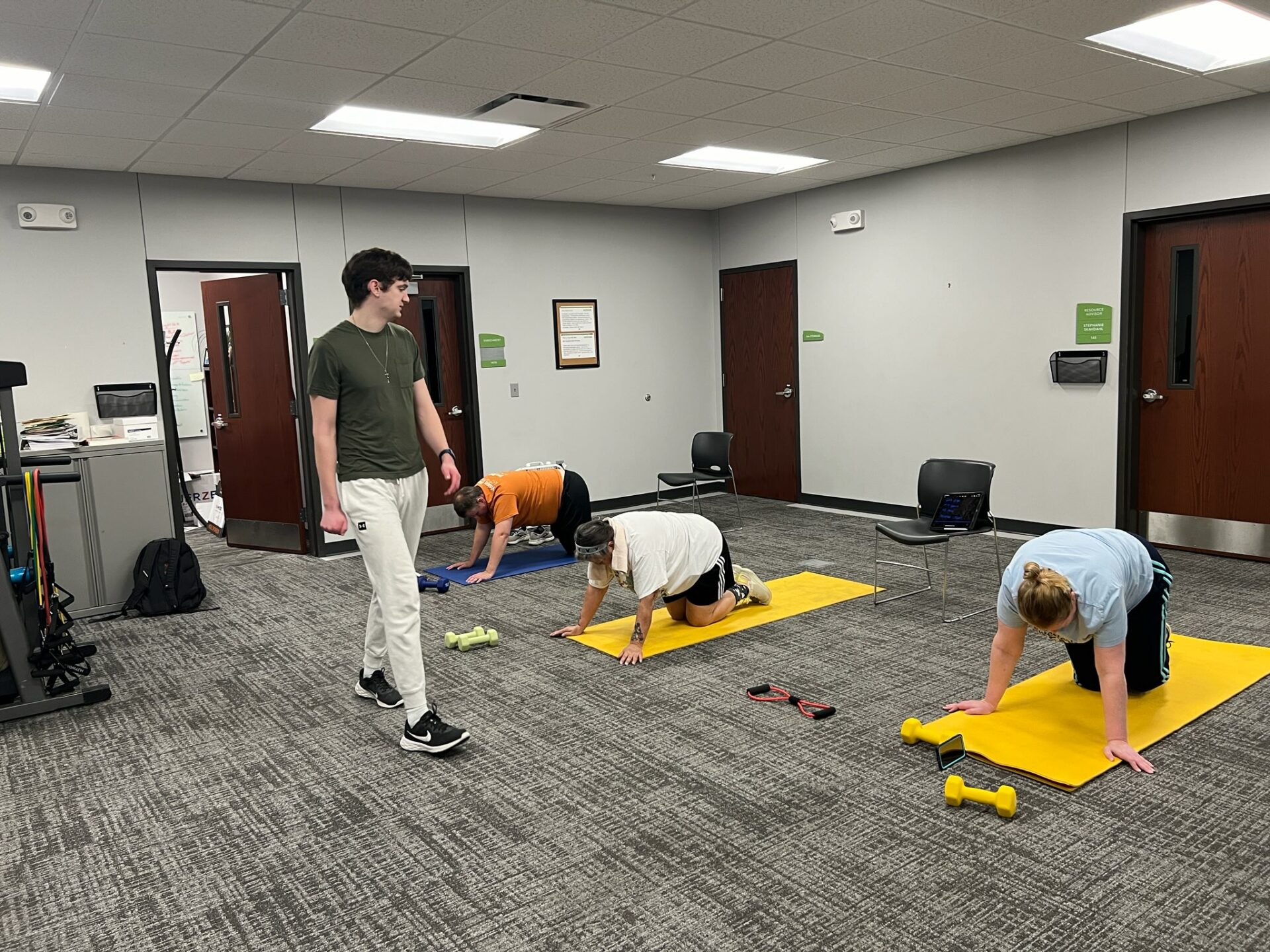 Isaac, the class instructor, at the front of the room describing the next workout move. Mickie, Francie and Kristal are doing the workout on yoga mats. For this move they are all positioned in a table top pose on their hands and knees. There are weights positioned near them.