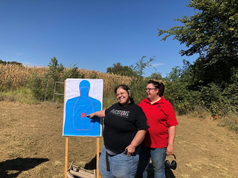 A target with the figure of a person on it. Willa is standing next to the target pointing at the hole she shot right in the middle of the bullseye. The instructor from the shooting range is standing next to her. There are blue skies and corn fields in the background.