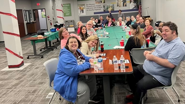 A group of about 25 adults sittting around one big table with Braille and Large print bingo cards.