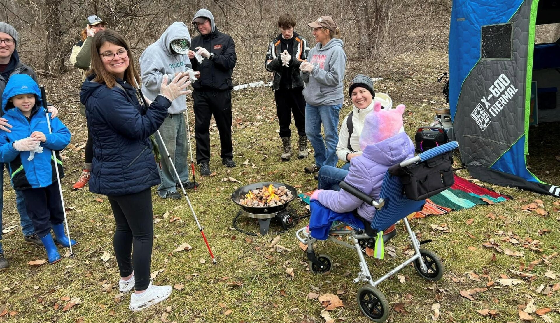 A group of about 8 people, a mix of teens and adults, standing around a campfire in the woods of Lake Cunningham.