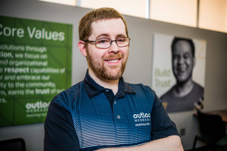 Aaron Peterson smiles for the camera while standing in front of an Outlook Nebraska Core Values poster