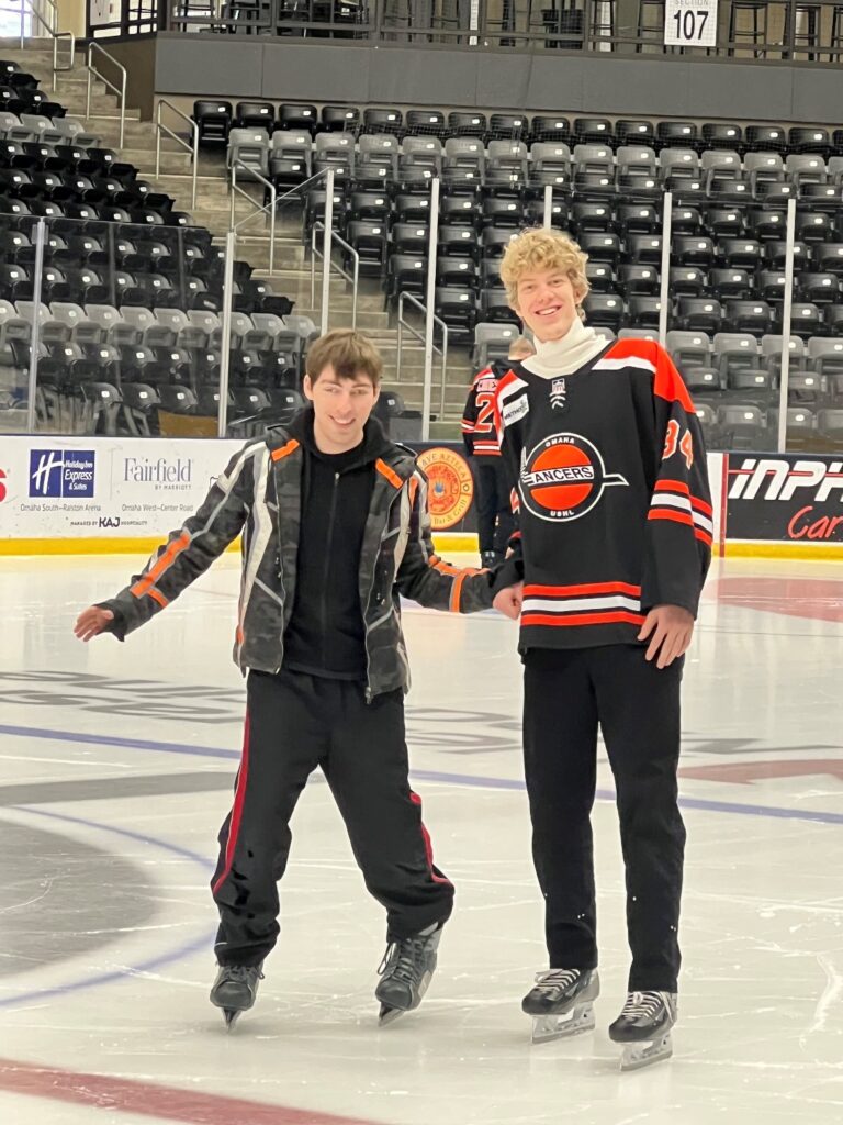 A lancers hockey player and a teenage boy ice skating on the hockey rink. The teenager is using the hockey players arm for support. they are both smiling and you can see stadium seating in the background.