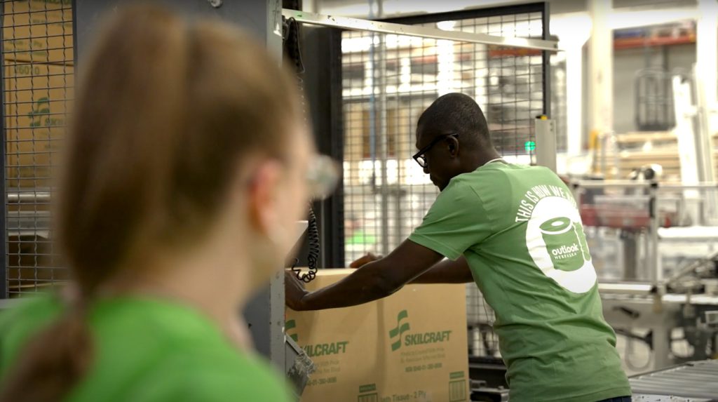 A man in a green shirt folds a SKillcraft cardboard box