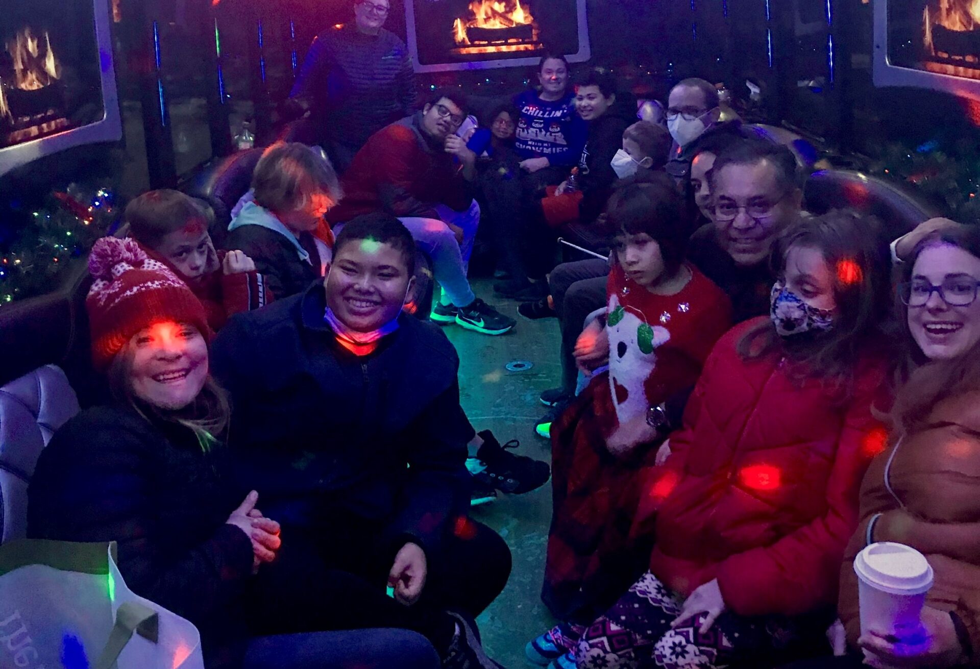 A group of teens in a shuttle on a holiday lights tour. The shuttle is lit up with llights on the ceiling and around the seats.