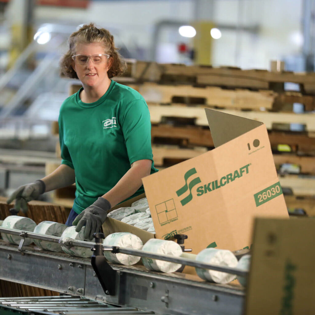 A woman in a green shirt works on the production line, a Skillcraft cardboard box sits next to her ready to be filled