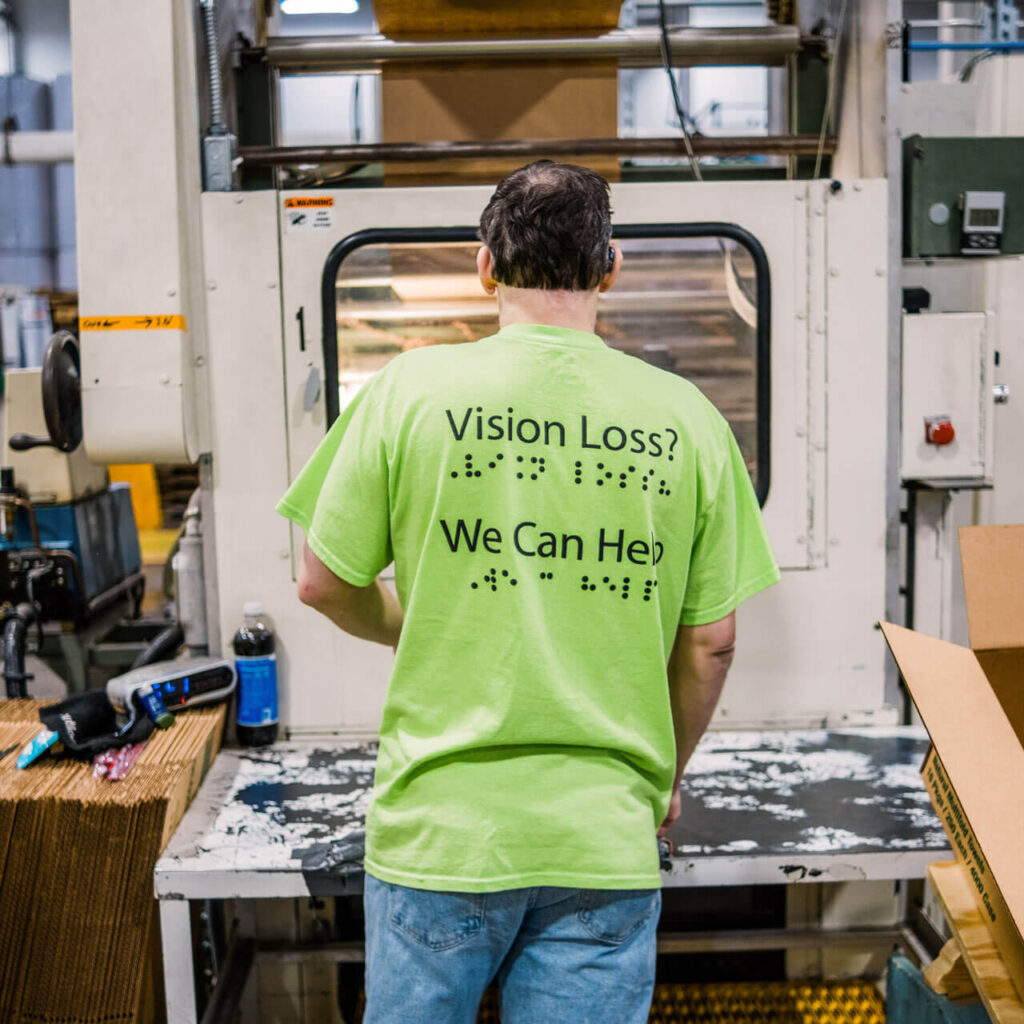 Back of a man working while wearing a green shirt that reads Vision Loss? We Can Help