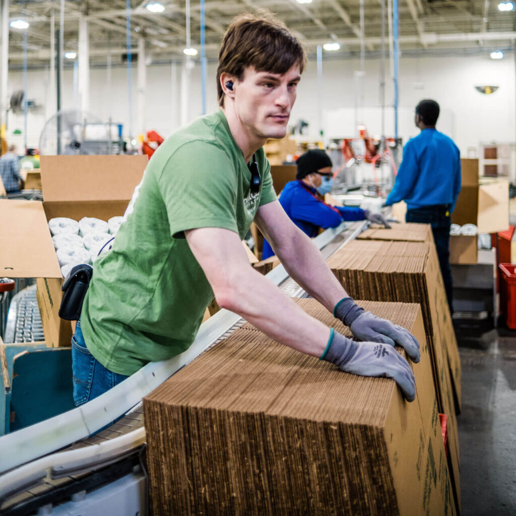 A man wearing gloves stacks cardboard boxes while two other employees work on the production line