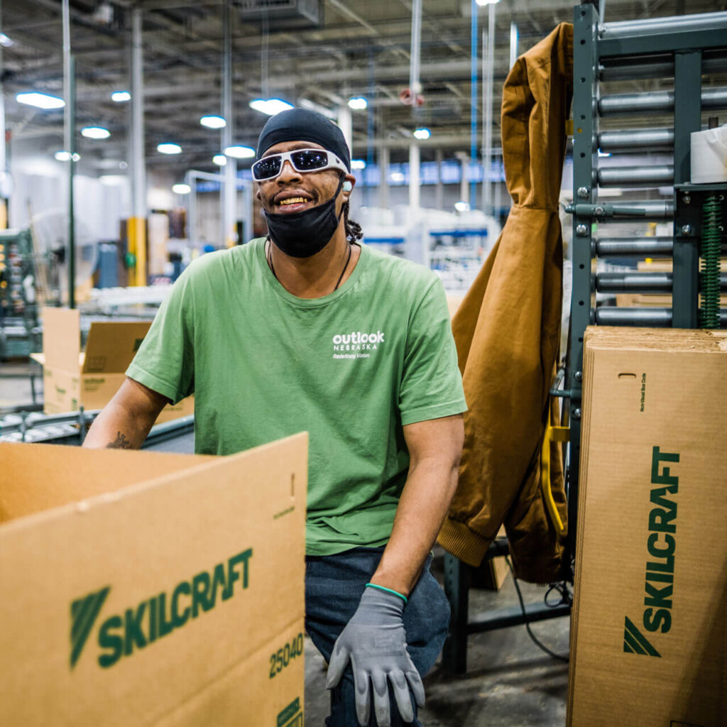 A man in a green Outlook Nebraska shirt sits next to several cardboard Skillcraft boxes