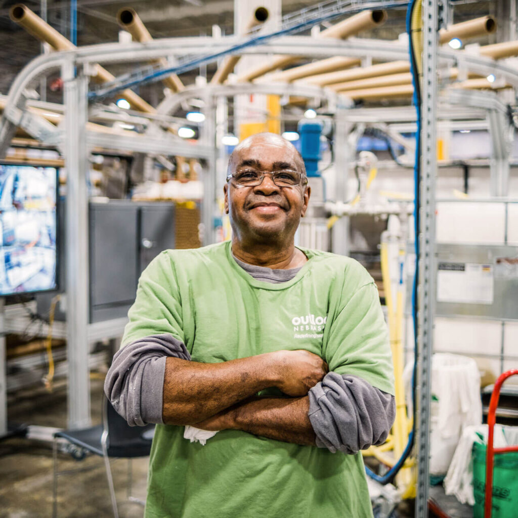 A man in a green shirt stands with his arms crossed in front of production equipment