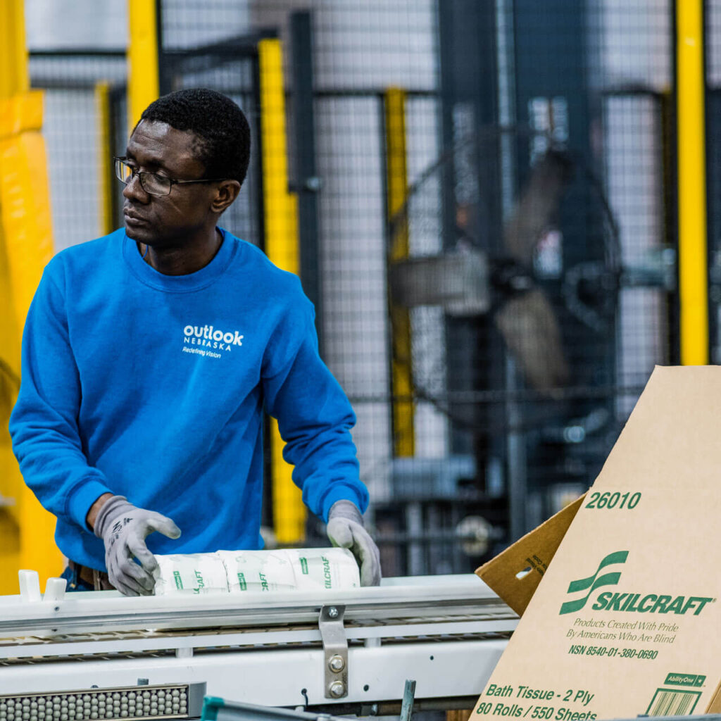 A man moves toilet paper along the production line. A Skillcraft cardboard box sits in the foreground