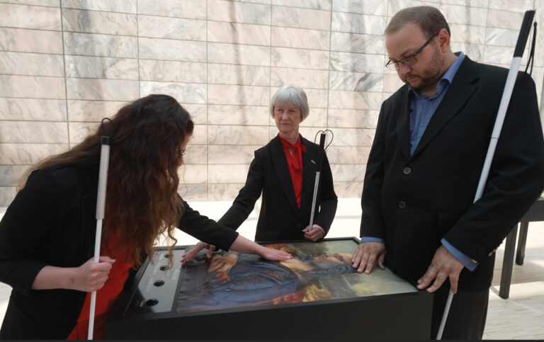 A picture provided by Joslyn Art Museum of two people standing on one side of a table. They each have a white can in one hand and are using their other hand to touch an art display. There is also someone on the other side of the table assisting them.