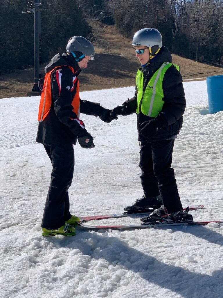 Two people facing eachother on a hill of snow. They are wearing skis and helmets. They are each extending a hand out to eachother