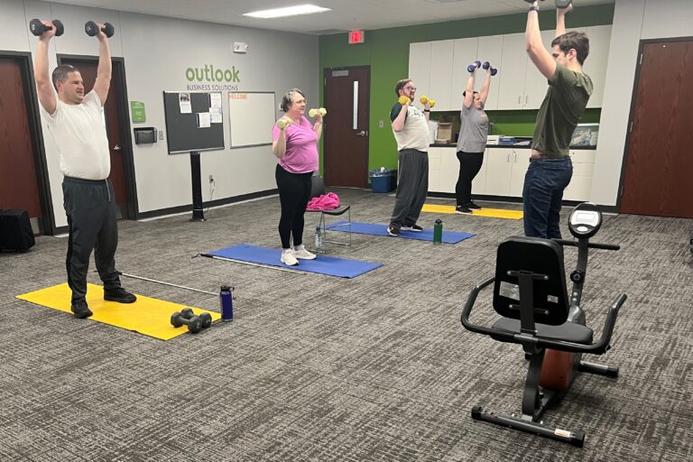 A line of 4 men and women holding dumb bells in the air during an exercise class. Isaac, the instructor, is at the front of the room giving directions.