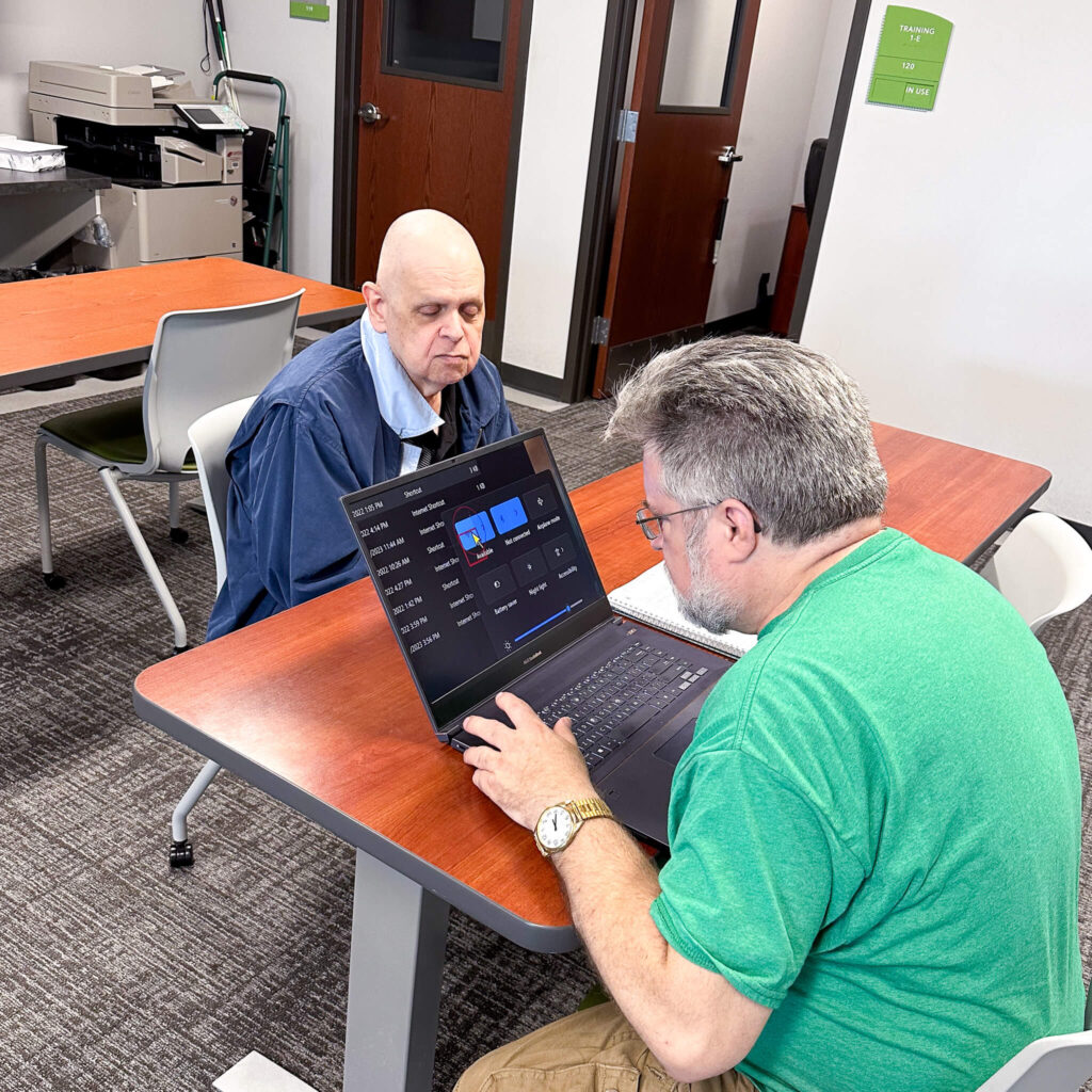 Man in a green shirt looks at a computer while another man looks on from across the table