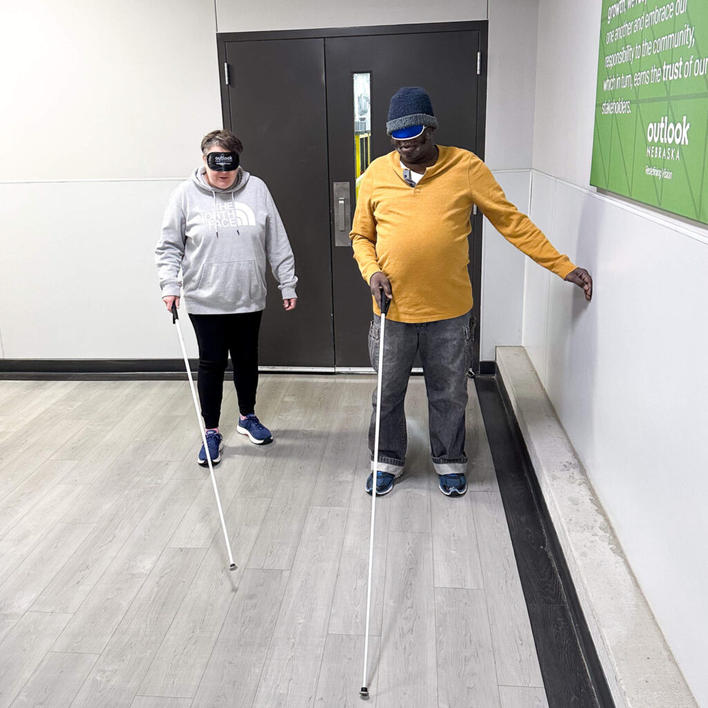 A white visually impaired woman walking down a hallway with a cane next to an African American man who also walks with a cane