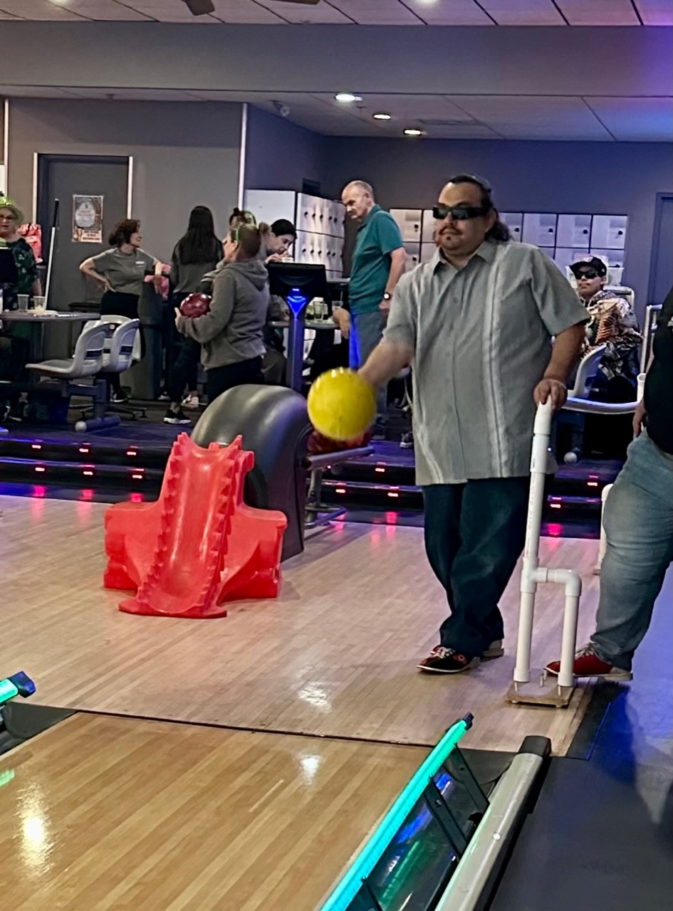 A man in a bowling alley wearing sunglasses. He has one hand on a white railing that leads up to the bowling lane. In the other hand he is swinging a bowling ball.