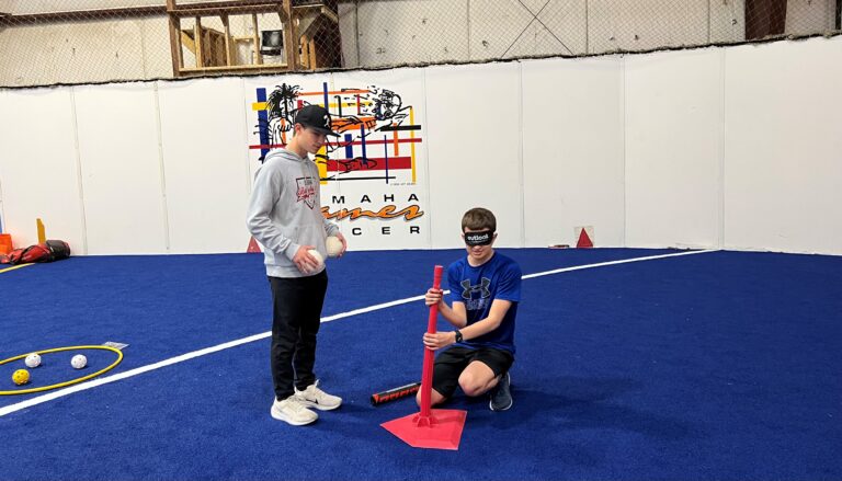 A young man is wearing sleep shades and kneeling down using both of his hands to figure out the height of the baseball tee. Another boy is standing next to him with a beep baseball in his hand.