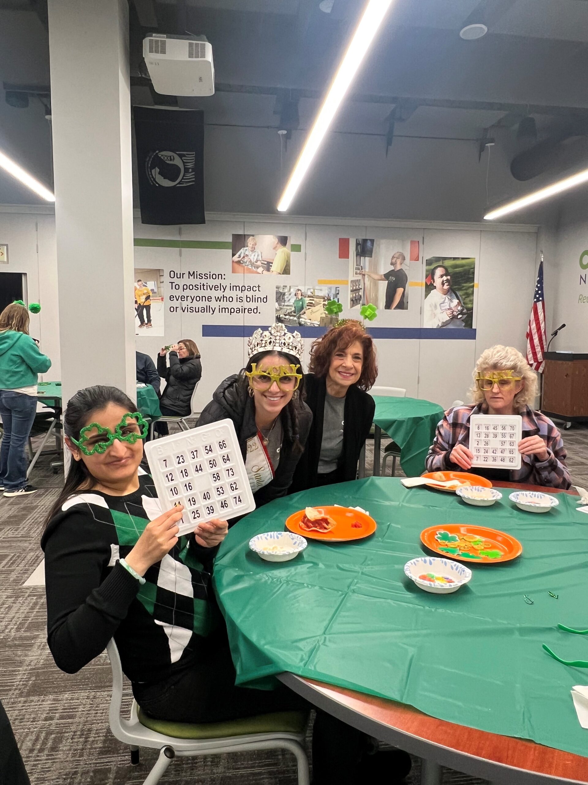 A table of three ladies wearing st. Patricks day themed glasses and headbands are sitting at a table. Two of them, Rachna and Melissa are holding up Bingo cards.