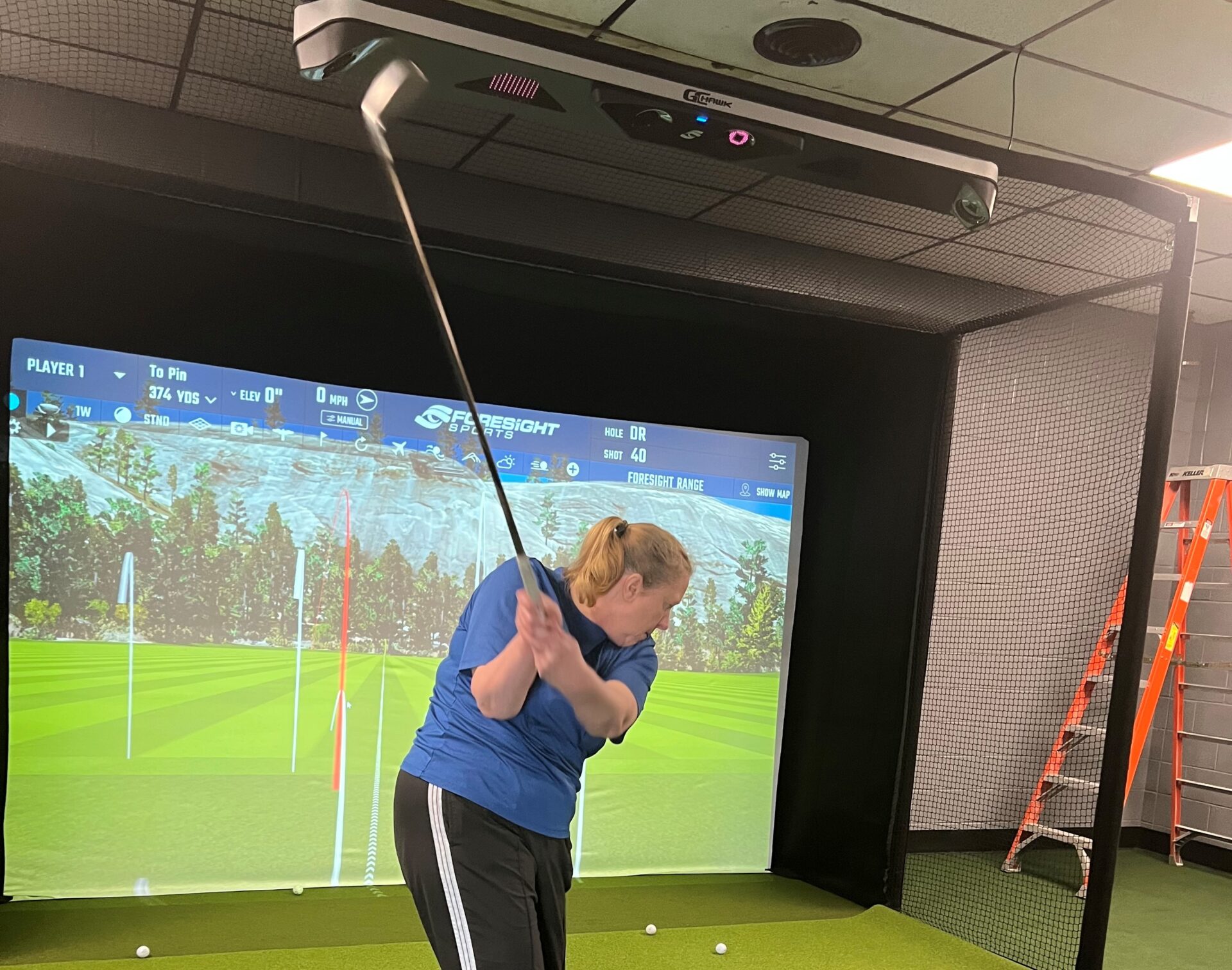 A woman in the full swing position with a golf club. There is a huge golf simulator screen in the background with views of a green golf course.