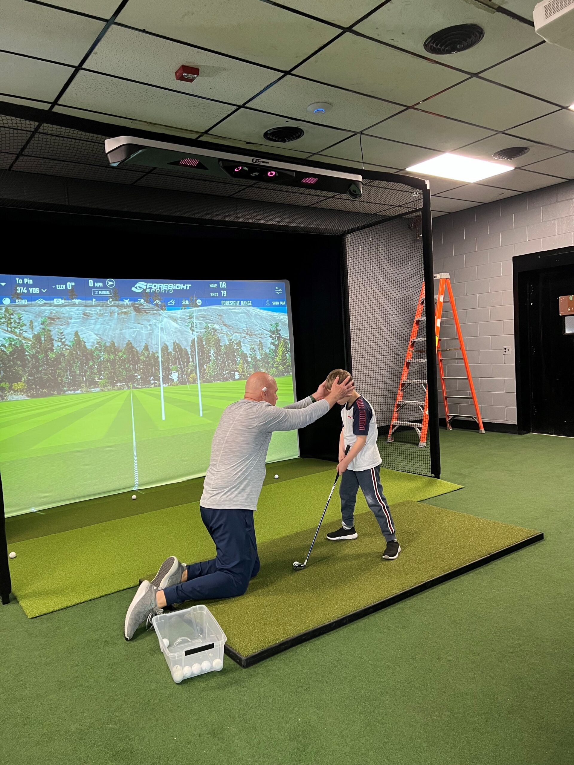 A young man with a golf club positioned in front of a golf simulator screen showing a picture of a golf course. There is an adaptive sports specialist is kneeling down in front of the young man and has his hands on either side of the young mans head to demonstrate how to swing the golf club properly without moving his head.