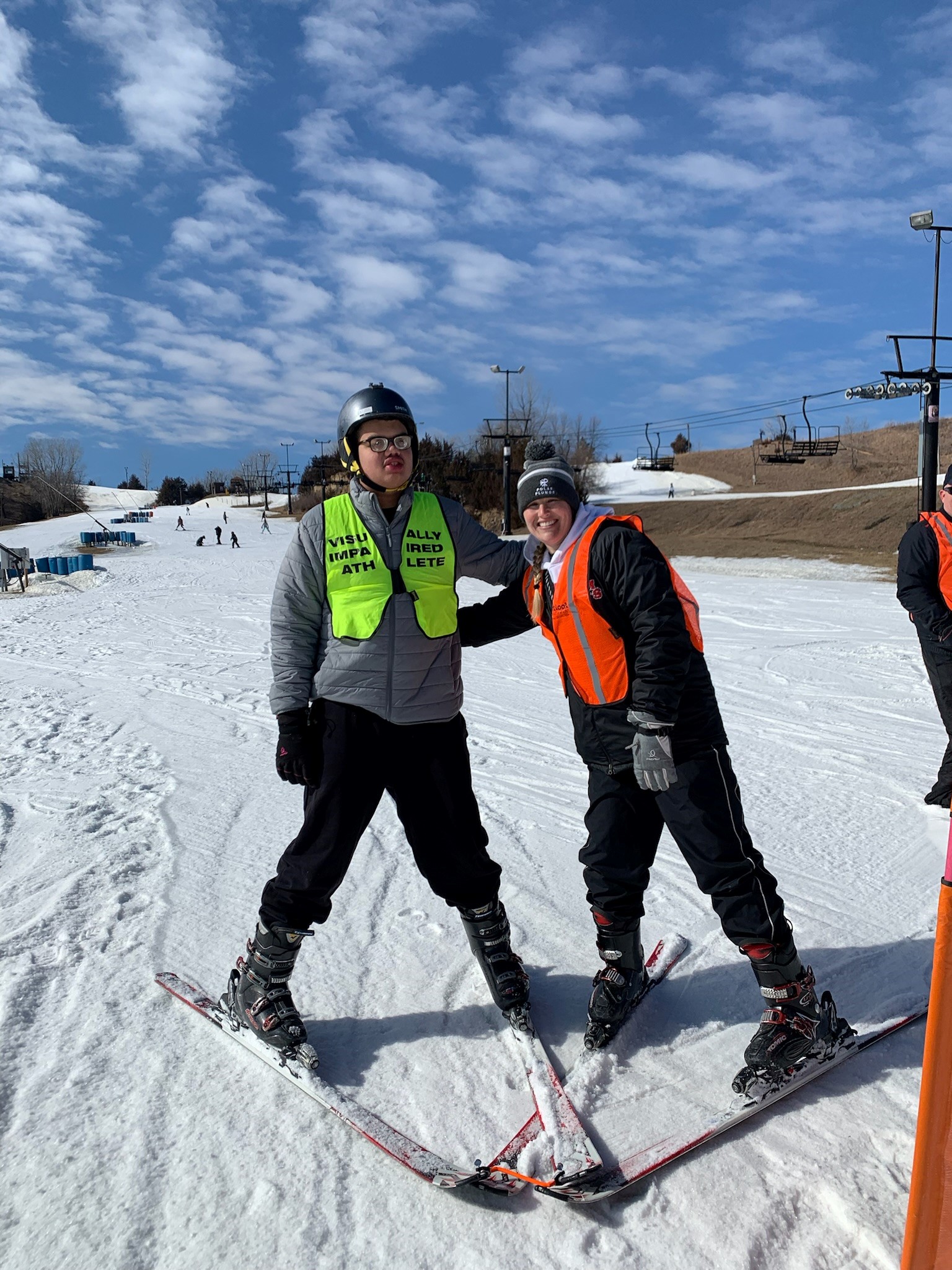 Two people wearing snow skis and snow gear are next to each other with their arms reaching out towards each other. There is a snow-covered hill, trees and blue sky in the background.