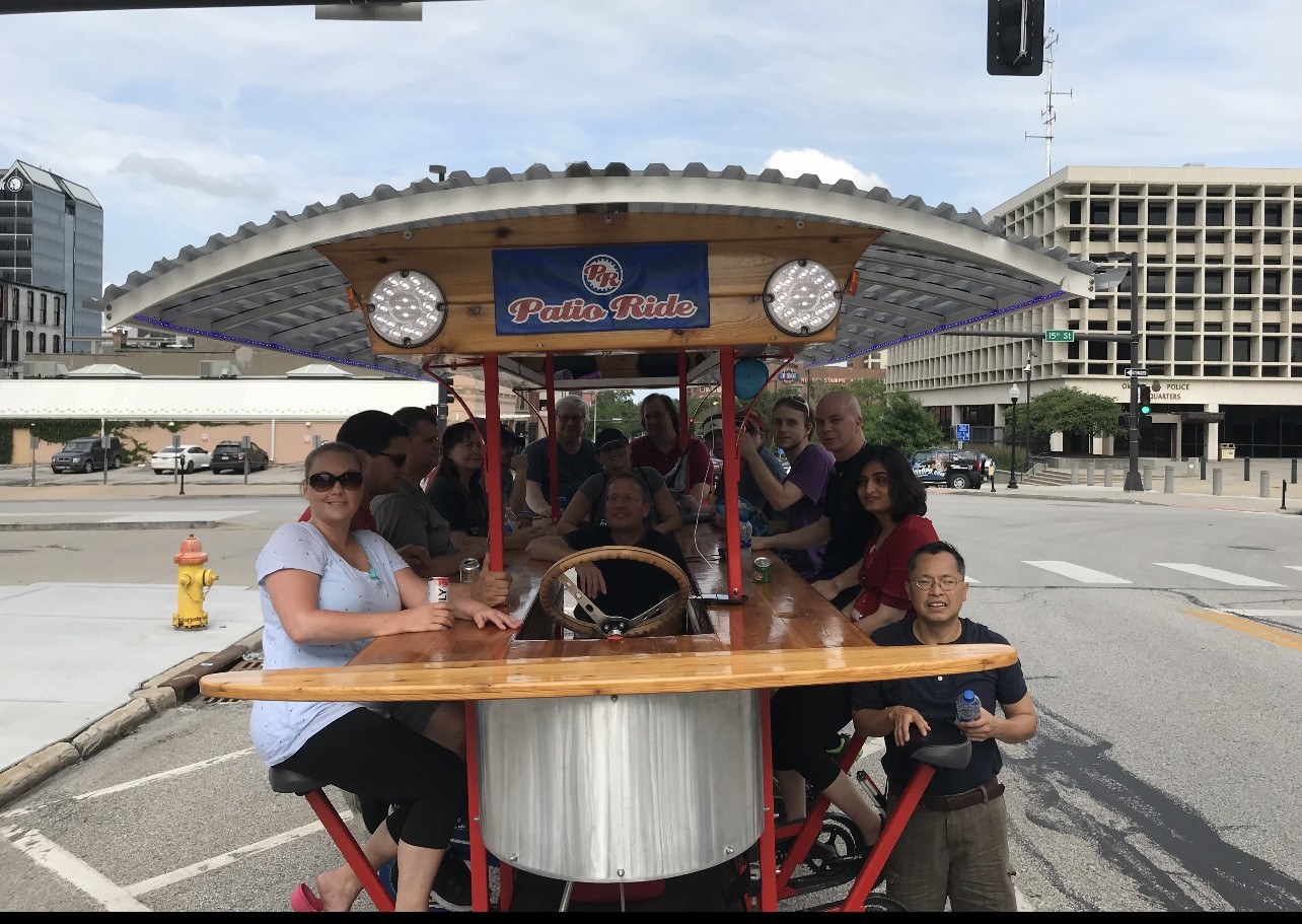 A shot from the front of the patio bike. It is like a trolley with bicycle seats along the sides. There is a group of 16 riders from Outlook sitting on the bike. There is a roof over the top of the bike.