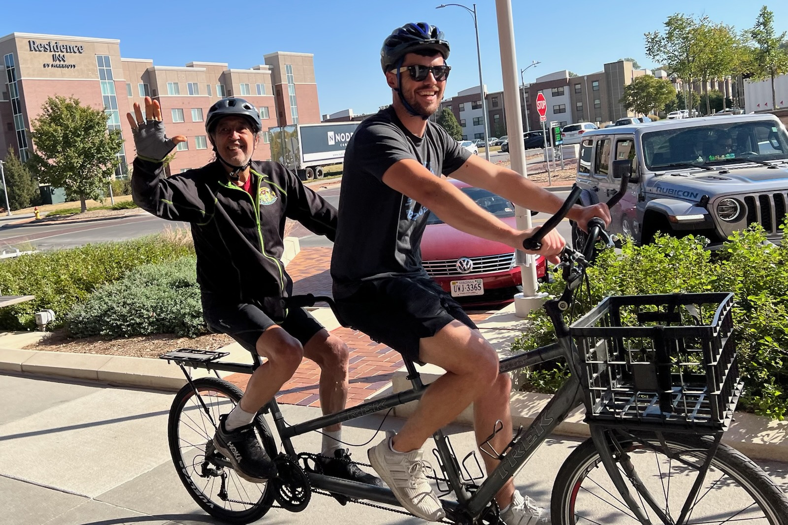 Two guys riding on a tandem bike. They are both smiling and the back seat rider is waving