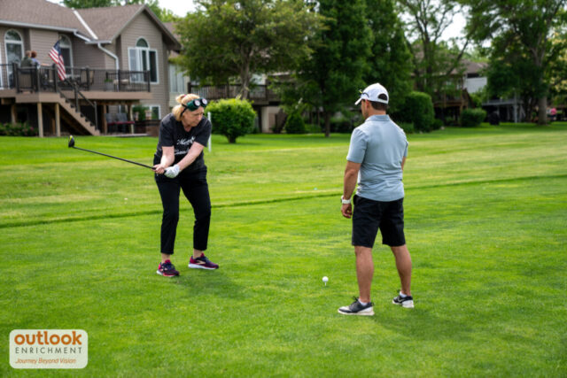A woman swinging with vision simulation goggles on her head while listening to instruction from a man.