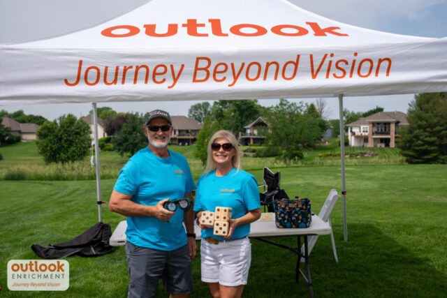 1 man and 1 woman volunteer smiling in front of their tent holding dice and vision simulation goggles.