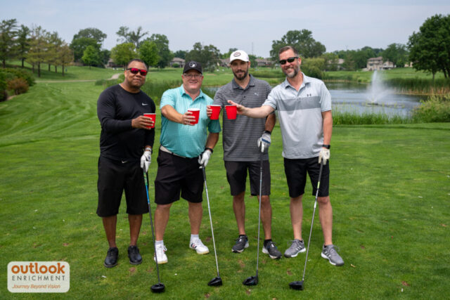 4 men smiling on the course and doing a cheers with their drinks.