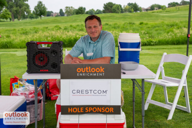 Crestcom sponsor hole. A man sitting at a table under a tent smiling.