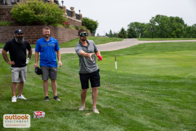 A group playing the cornhole challenge. A man is throwing a bag with vision simulation goggles on.