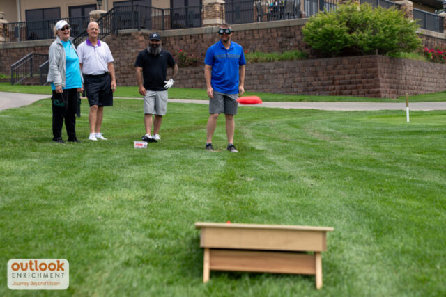 A group playing the cornhole challenge. A man is throwing a bag with vision simulation goggles on.