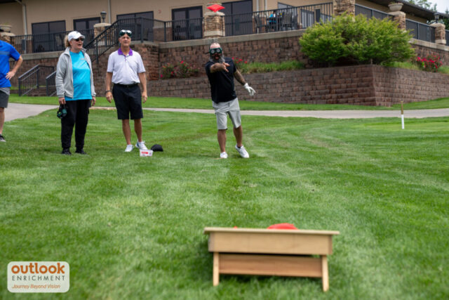 A group playing the cornhole challenge. A man is throwing a bag with vision simulation goggles on.