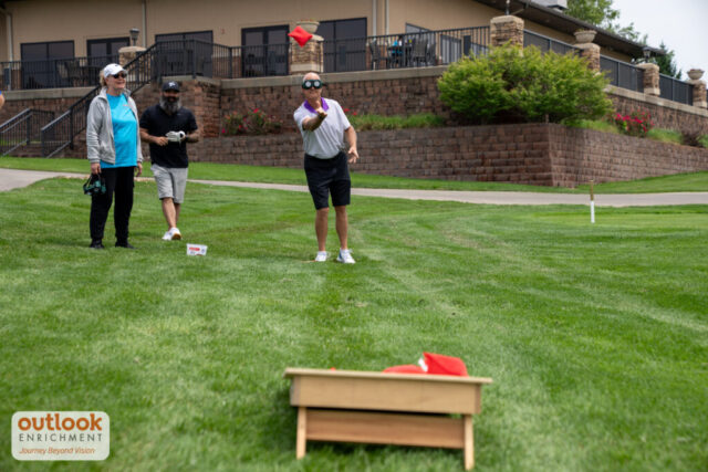 A group playing the cornhole challenge. A man is throwing a bag with vision simulation goggles on.