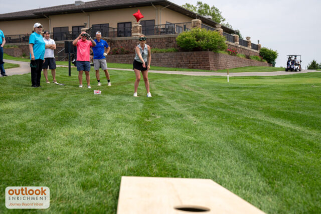 A group playing the cornhole challenge. A woman is throwing a bag with vision simulation goggles on.