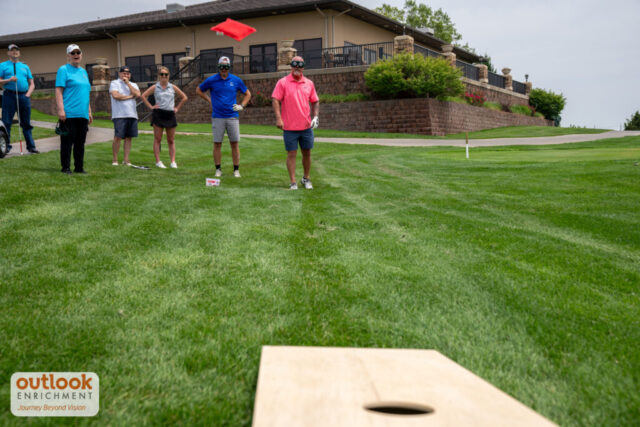 A group playing the cornhole challenge. A man is throwing a bag with vision simulation goggles on.