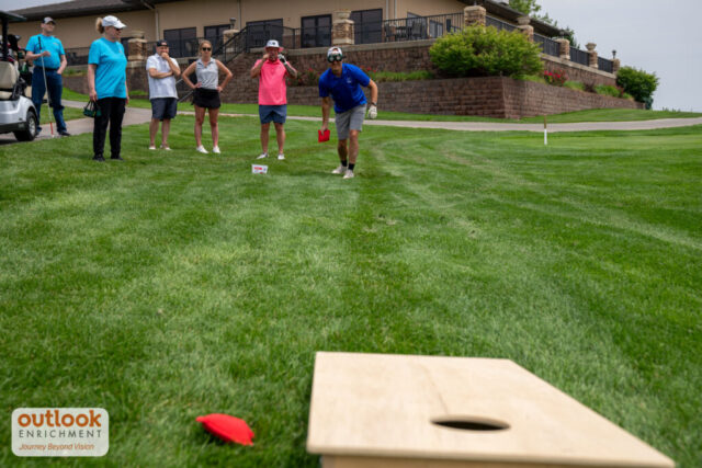 A group playing the cornhole challenge. A man is throwing a bag with vision simulation goggles on.