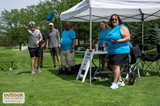 Group watching as a man does cornhole with vision simulation goggles on.