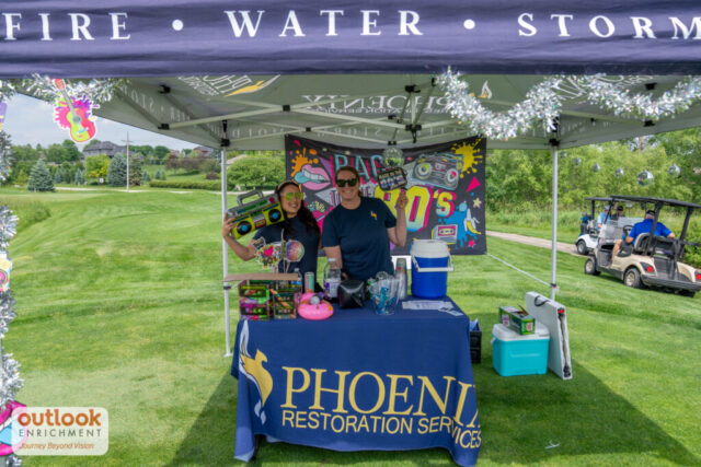 Phoenix Restoration Services sponsor hole. 2 ladies holding up 80's themed props.