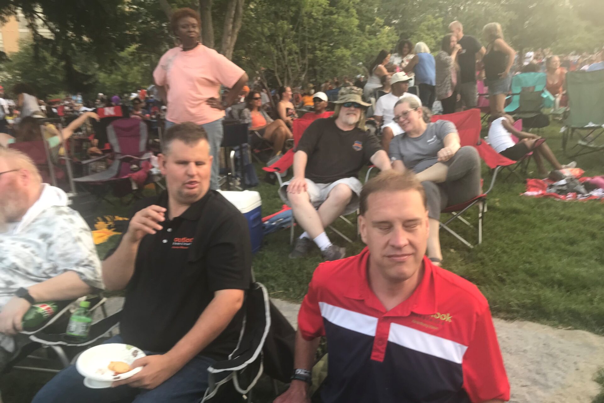 Mickie and Donovan at a large grassy park sitting in lawn chairs an, listening to music and eating snacks. In the background there are lots of other people in lawn chairs and sitting on blankets.
