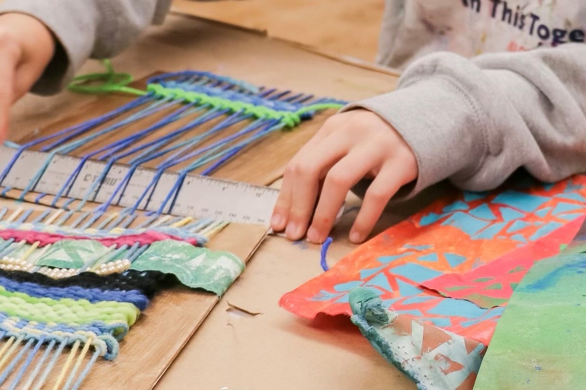 Hands working with a waving loom. There is a partial weaving project in progress and other weaving materials on the surface.