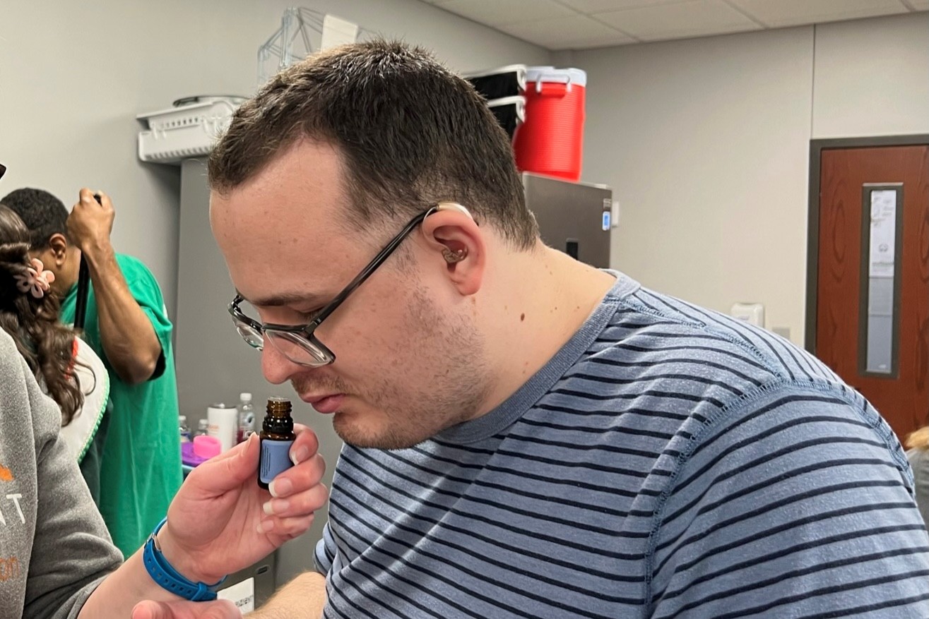 Alek is wearing thick glasses and a striped shirt. Someone is holding up a little glass jar of scented oil up towards his nose for hime to smell.