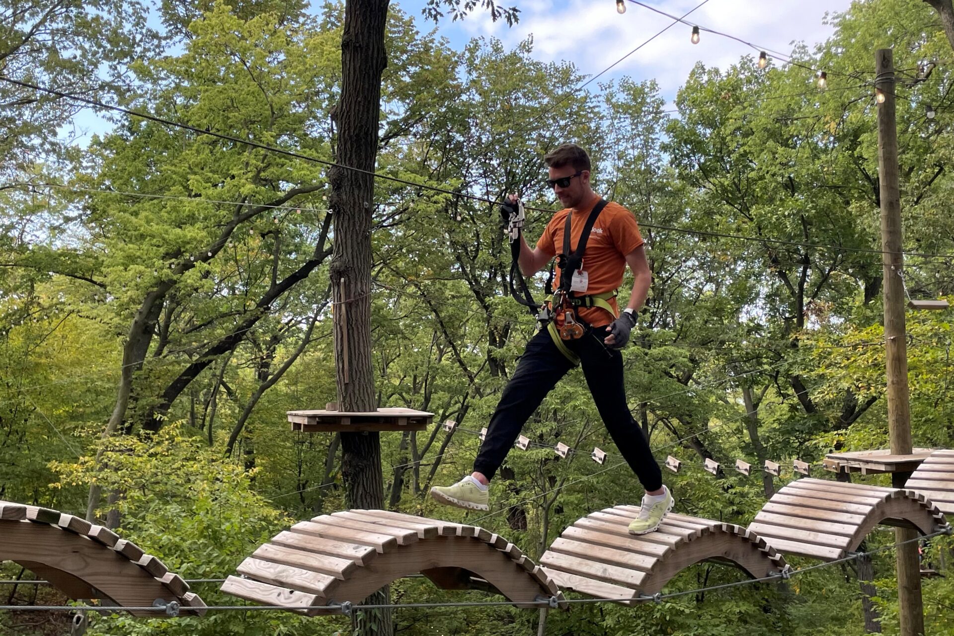 A man wearing sunglasses is leaping across a wavy wooden bridge that is suspended in the air at TreeRush Adventures. There are trees in the background.